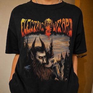 May include: A black short-sleeve t-shirt featuring a graphic print with the text "ELECTRIC WIZARD" in orange and yellow flames. The design includes a bearded, horned figure with glowing eyes, smoke, and a dark castle silhouette in the background.