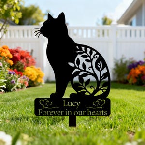 May include: Black metal cat garden stake with a decorative leaf design on the cat's body. The stake includes a rectangular sign that reads "Lucy Forever in our hearts". The cat is sitting on a green lawn.