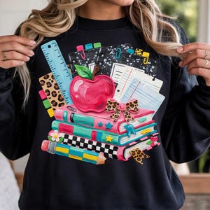Peut inclure: Un sweat-shirt noir à manches longues avec une impression graphique de fournitures scolaires, notamment une pile de livres, une pomme rouge, une règle bleue, un crayon et un nœud à motif léopard. Le design inclut divers accents colorés.