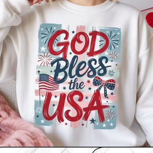Peut inclure: Sweat-shirt blanc avec un motif patriotique. Le graphisme présente le texte "GOD Bless the USA" en rouge et bleu, ainsi qu'un drapeau américain, des feux d'artifice et des étoiles. Le motif est encadré de rectangles bleus et roses.