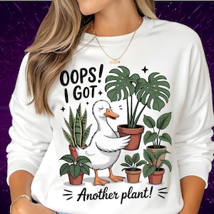 Peut inclure: Sweat-shirt blanc avec un dessin d'un canard blanc tenant une plante en pot, entouré d'autres plantes en pot. Le texte dit "OOPS! I GOT + Another plant!" en noir. Le canard a un bec et des pattes oranges. Les plantes sont vertes et dans des pots bruns.