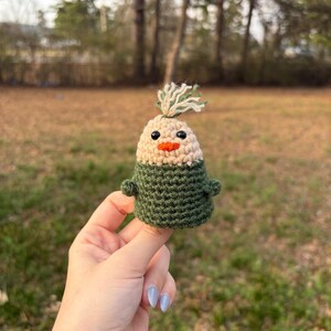 May include: A handmade crochet chick with a beige head, green body, and orange beak. The chick has small black eyes and a tuft of green and white yarn on top. The chick is held up by a person's fingers.