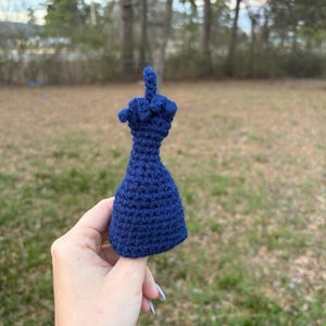 May include: A navy blue crocheted middle finger, a novelty item. The handmade piece is held up against a blurred outdoor background of trees and grass. The crocheted item is a unique, handcrafted gift.