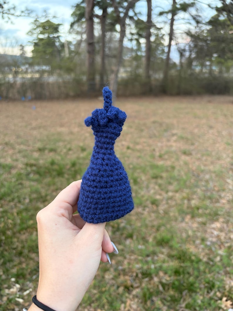 May include: A navy blue crocheted middle finger, a novelty item. The handmade piece is held up against a blurred outdoor background of trees and grass. The crocheted item is a unique, handcrafted gift.