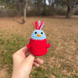May include: A handmade crochet bird figurine. The bird has a light blue head with a red and white tuft, a red body, and small red wings. The bird has a yellow beak and black eyes. The figurine is held in a hand against a blurred outdoor background.