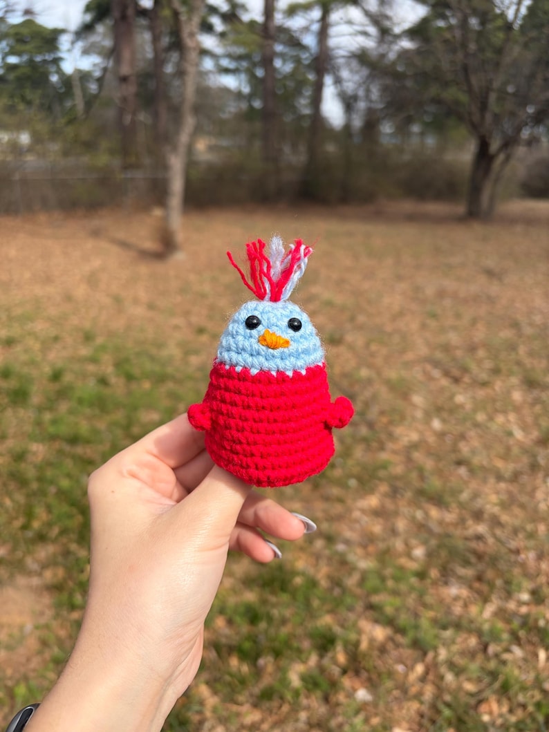 May include: A handmade crochet bird figurine. The bird has a light blue head with a red and white tuft, a red body, and small red wings. The bird has a yellow beak and black eyes. The figurine is held in a hand against a blurred outdoor background.