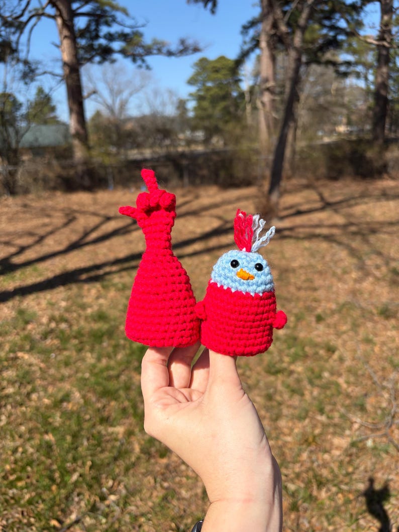 May include: Two handmade crochet figures. One is a red hand with the middle finger raised. The other is a blue and red bird with a yellow beak and white and blue accents. The figures are held up against a blurred outdoor background.