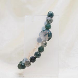 May include: A beaded bracelet featuring a mix of green and gray round stones. The bracelet includes a large, spherical stone with a unique pattern of green and white, and smaller, solid green beads. The bracelet is displayed on a clear stand.