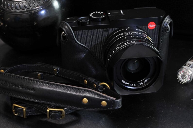 May include: A black camera with a large lens and a black leather strap. The lens has the words 'SUMMILUX 1:1.7/28 ASPH' printed on it. The camera has a black leather case with a gold buckle.