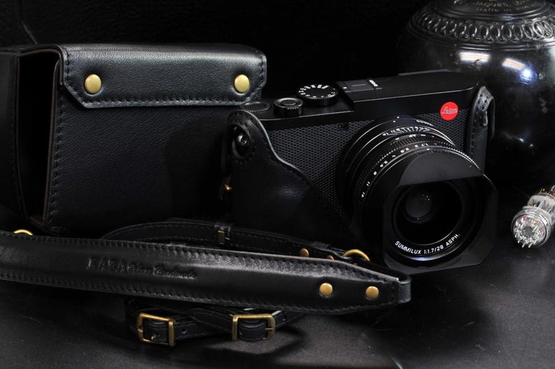 May include: A black leather camera strap with gold accents and a black camera with a lens and a red dot on the top.
