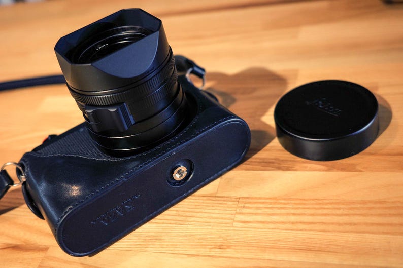 May include: A black camera lens with a black lens hood attached. The lens is in a black leather camera case. A black lens cap is on a wooden surface next to the camera.