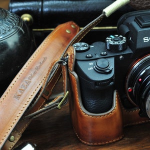Peut inclure: Un appareil photo Sony noir avec un étui et une sangle en cuir marron. L'appareil photo a un objectif argenté avec le texte "Voigtlander" dessus. La sangle est étiquetée "Kaza Delta Handmade".
