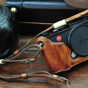 以下が含まれることがあります： 黒いカメラが入った茶色の革製のカメラケース。カメラには「Leica」と書かれたレンズキャップが付いています。ケースには白いストラップアジャスターが付いたストラップが付いています。