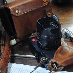 May include: A brown leather camera strap with a black camera lens attached. The camera lens has a black lens cap and a silver ring with numbers on it. The camera strap is attached to a brown leather camera case.
