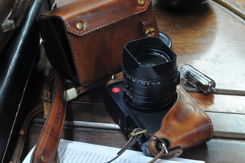 May include: A brown leather camera strap with a black camera lens attached. The camera lens has a black lens cap and a silver ring with numbers on it. The camera strap is attached to a brown leather camera case.