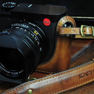 May include: A black and silver camera lens with a brown leather camera case. The lens has a black lens hood and a silver lens barrel with the text 'SUMMILUX 1:1.7/28 ASPH' printed on it. The camera case has a brown leather strap with the text 'KAZZA Leather Handmade' printed on it.