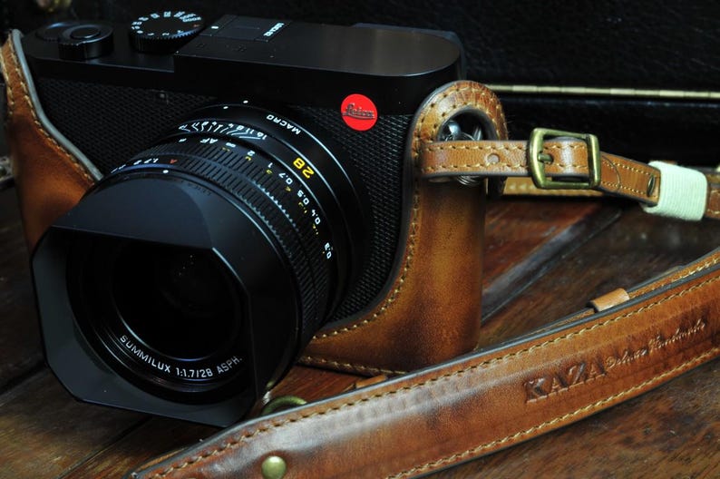 May include: A black and silver camera lens with a brown leather camera case. The lens has a black lens hood and a silver lens barrel with the text 'SUMMILUX 1:1.7/28 ASPH' printed on it. The camera case has a brown leather strap with the text 'KAZZA Leather Handmade' printed on it.