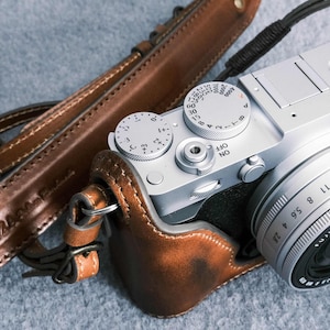 Könnte beinhalten: Silberne Fujifilm X-E3 Kamera mit Objektiv, brauner Lederhülle und passendem Riemen. Die Kamera hat Drehknöpfe und Tasten. Der Lederriemen ist mit dem Wort "RAZA" geprägt.