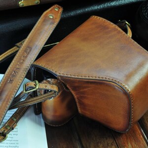 May include: A brown leather camera bag with a strap that says 'KAYA LABS' on it. The bag has a flap that closes with a buckle. The bag is sitting on a wooden surface.