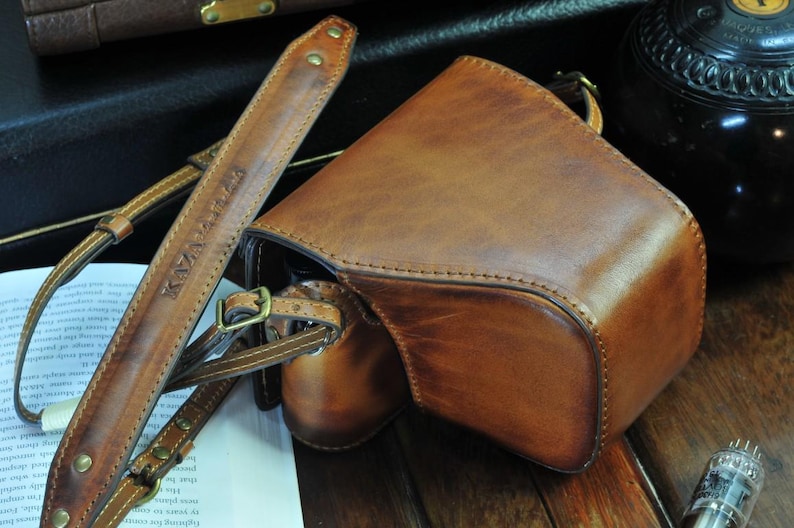 May include: A brown leather camera bag with a strap that says 'KAYA LABS' on it. The bag has a flap that closes with a buckle. The bag is sitting on a wooden surface.