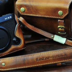 Könnte beinhalten: Eine braune Lederkamera-Tasche mit einem Riemen und dem eingeprägten Text "KAZAN" auf dem Riemen. Die Tasche ist für eine Fujifilm X-E3 Kamera konzipiert.