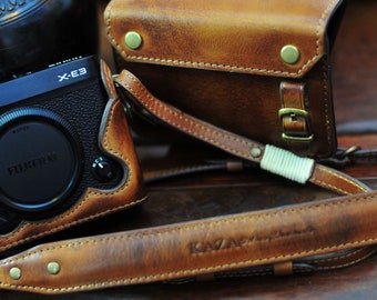 May include: A brown leather camera case with a strap and the text "KAZAN" embossed on the strap. The case is designed for a Fujifilm X-E3 camera.