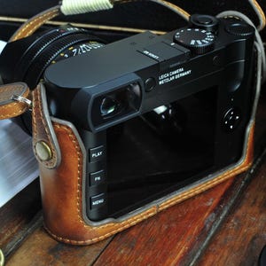May include: A black Leica camera with a brown leather case. The camera has a viewfinder, a lens, and a series of buttons on the side. The text on the camera reads 'Leica Camera Wetzlar Germany'.