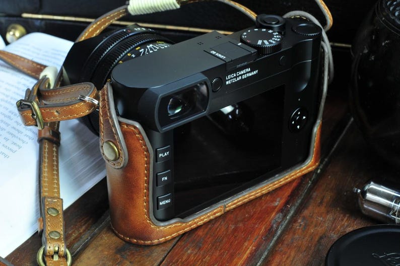 May include: A black Leica camera with a brown leather case. The camera has a viewfinder, a lens, and a series of buttons on the side. The text on the camera reads 'Leica Camera Wetzlar Germany'.