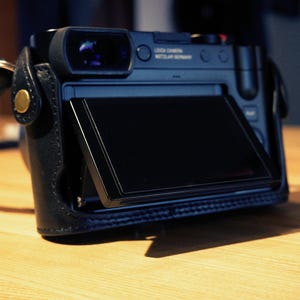 May include: A black digital camera with a flip-up screen. The camera has a leather case and a gold accent on the front. The screen is displaying a black screen.