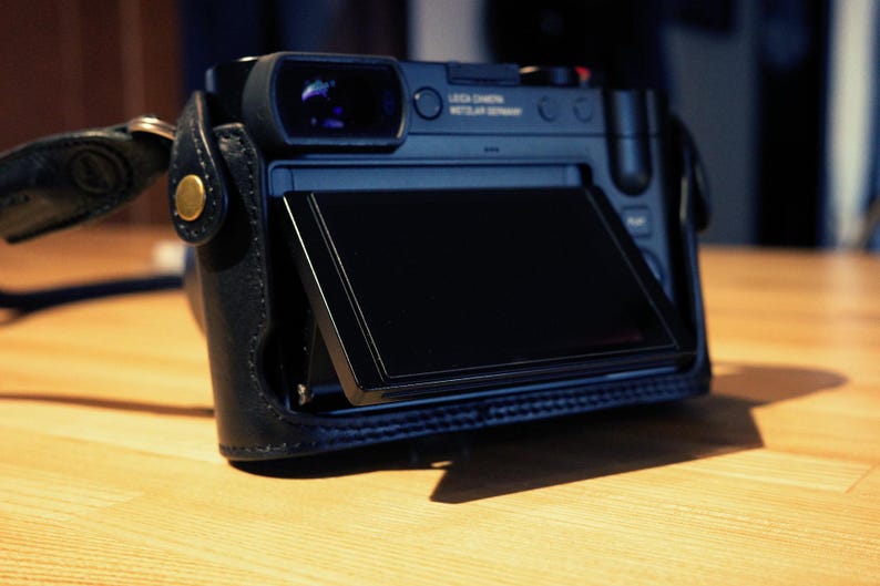 May include: A black digital camera with a flip-up screen. The camera has a leather case and a gold accent on the front. The screen is displaying a black screen.