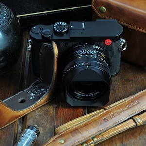 May include: A black camera with a brown leather camera strap and a brown leather camera case. The camera has a large lens and a red button on the top. The case has the words 'Limited KAZA' printed on it. The strap has the words 'KAZAK Leather' printed on it.
