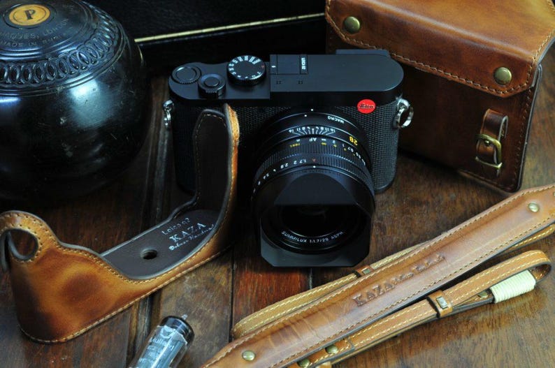May include: A black camera with a brown leather camera strap and a brown leather camera case. The camera has a large lens and a red button on the top. The case has the words 'Limited KAZA' printed on it. The strap has the words 'KAZAK Leather' printed on it.