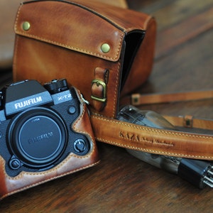 Pode incluir: Uma câmera Fujifilm X-T2 preta com uma capa e alça de couro marrom. A câmera está em uma capa de couro marrom, e a alça também é de couro marrom. A capa e a alça têm um visual vintage.