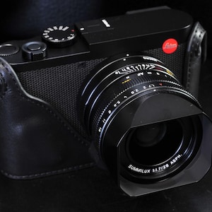 May include: A black camera with a black leather case. The camera has a silver lens with the words 'SUMMILUX 1:1.7/28 ASPH.' printed on it. The lens has a black lens hood.