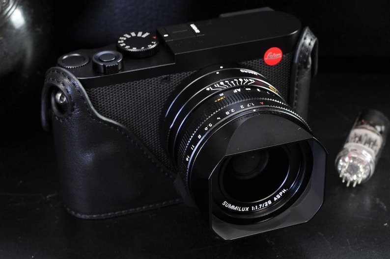 May include: A black camera with a black leather case. The camera has a silver lens with the words 'SUMMILUX 1:1.7/28 ASPH.' printed on it. The lens has a black lens hood.