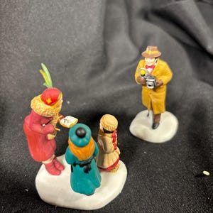 May include: A miniature Christmas village scene featuring four figurines. One figure in a yellow coat and hat holds a lantern. Three figures are grouped together, one in a red coat, one in teal, and one in a tan coat.