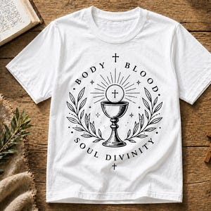 May include: White t-shirt featuring a black graphic design with the words "BODY BLOOD SOUL DIVINITY" surrounding a chalice and host. The design includes a cross and laurel wreath. The shirt is displayed on a wooden surface.