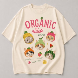 Może przedstawiać: Kremowa koszulka z nadrukiem przedstawiającym kreskówkowe koty w owocowych kapeluszach. Nad kotami widnieje napis "ORGANIC Sweet Crews", a pod nimi "Sweetness Straight From Me". Projekt zawiera ananasa, truskawkę i arbuza.