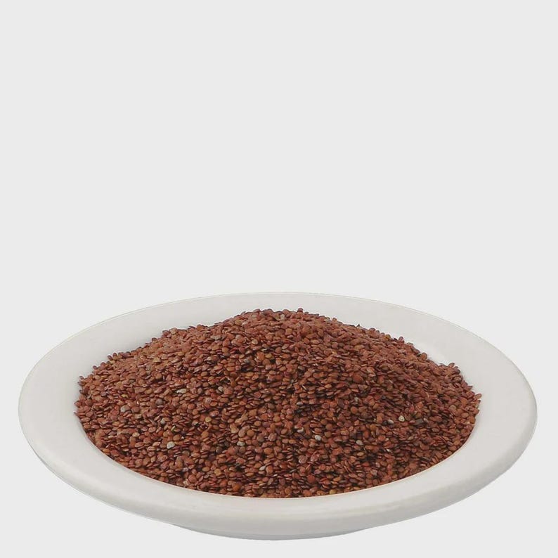 May include: A mound of small, reddish-brown seeds piled in the center of a plain white ceramic plate. The seeds have a smooth, oval shape and a consistent dark brown color, creating a textured heap that sits neatly within the circular rim.