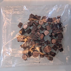 May include: A clear plastic bag filled with pieces of dark brown, chopped wood. The wood pieces vary in size and shape, with some appearing to be cross-sections of small branches. The bag is sealed and sits on a light-colored surface.