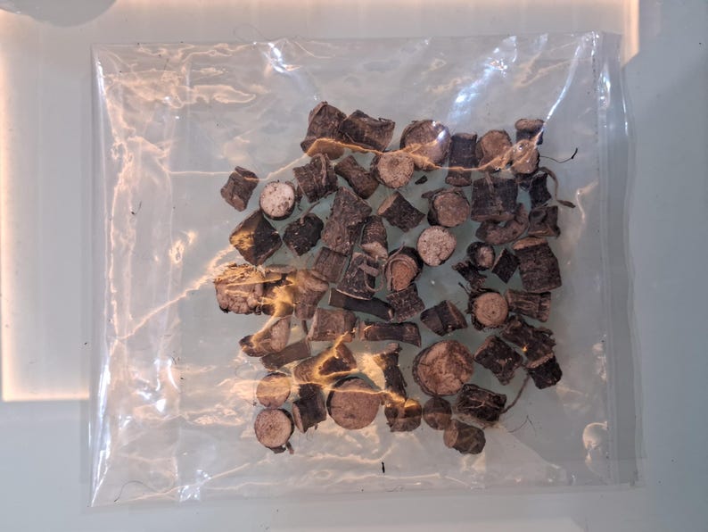 May include: A clear plastic bag filled with pieces of dark brown, chopped wood. The wood pieces vary in size and shape, with some appearing to be cross-sections of small branches. The bag is sealed and sits on a light-colored surface.