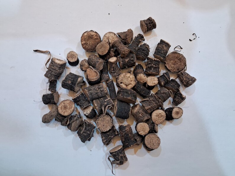 May include: A collection of dark brown, dried root pieces, varying in size and shape, scattered on a white surface. Some pieces are cylindrical, while others are irregular, with visible textures and cut ends. The overall color palette is earthy.
