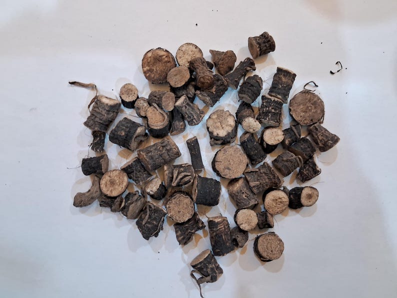 May include: A collection of small, dark brown, and tan pieces of wood, possibly roots or stems, scattered on a white surface. The pieces vary in shape, with some appearing cylindrical and others more irregular.
