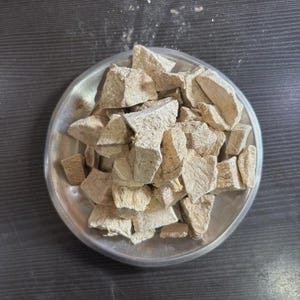 May include: A pile of irregular, light beige, dried root chunks resting on a round, metallic silver plate. The pieces have a rough, fibrous, and matte texture with varying shapes and sizes, appearing dry and brittle in their natural state.