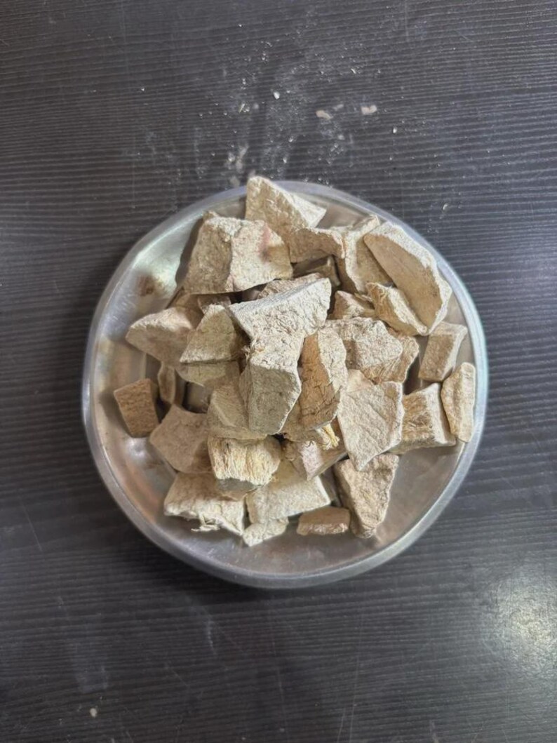 May include: A pile of irregular, light beige, dried root chunks resting on a round, metallic silver plate. The pieces have a rough, fibrous, and matte texture with varying shapes and sizes, appearing dry and brittle in their natural state.