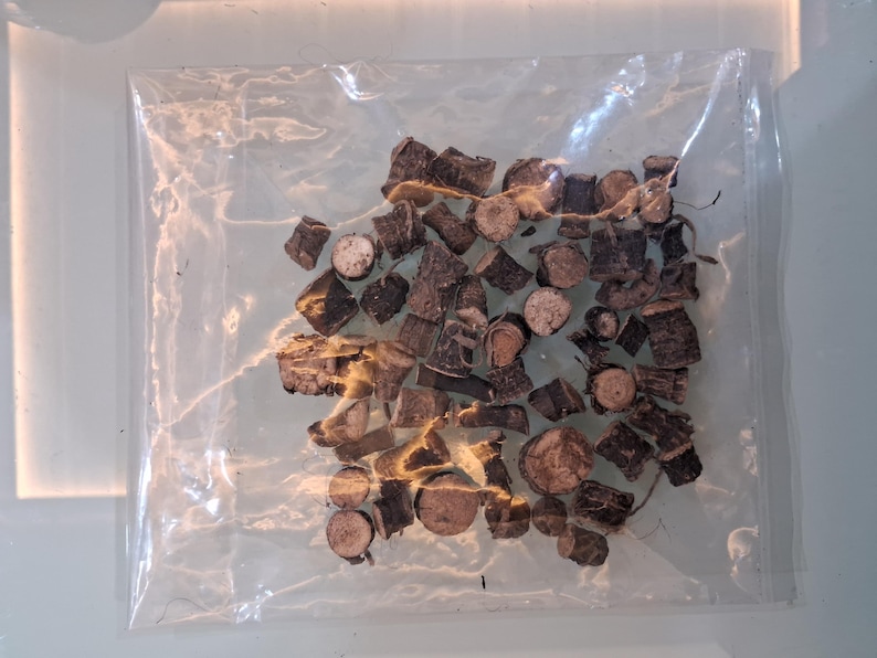 May include: A clear plastic bag filled with small, irregularly shaped pieces of dark brown wood. Some pieces appear to be cross-sections of branches, showing circular patterns. The wood pieces vary in size and shape, creating a textured appearance.