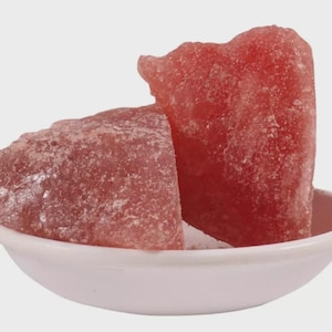 May include: Two large, rough-cut, pink Himalayan salt chunks rest in a small, white, shallow bowl. The salt crystals have a reddish hue and a textured surface, suggesting a natural, unrefined product. The bowl is simple and clean.