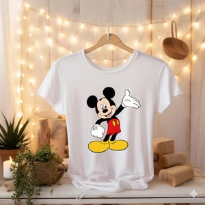 Puede incluir: Camiseta blanca con un dibujo de Mickey Mouse. El personaje de dibujos animados se muestra con orejas negras, cuerpo negro, pantalones cortos rojos, zapatos amarillos y guantes blancos. La camiseta est&aacute; colgada de una percha de madera.