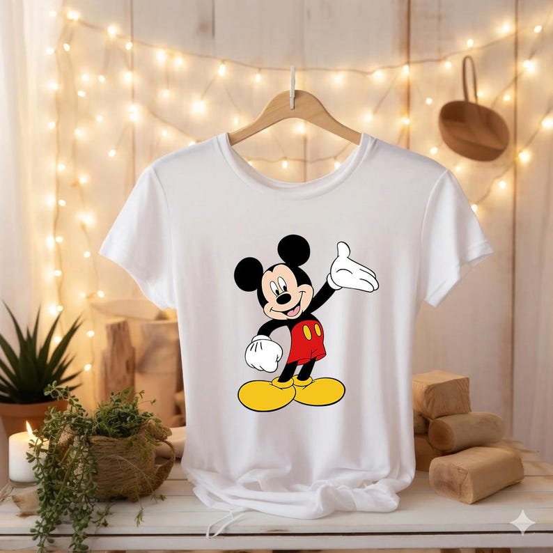 Puede incluir: Camiseta blanca con un dibujo de Mickey Mouse. El personaje de dibujos animados se muestra con orejas negras, cuerpo negro, pantalones cortos rojos, zapatos amarillos y guantes blancos. La camiseta est&aacute; colgada de una percha de madera.