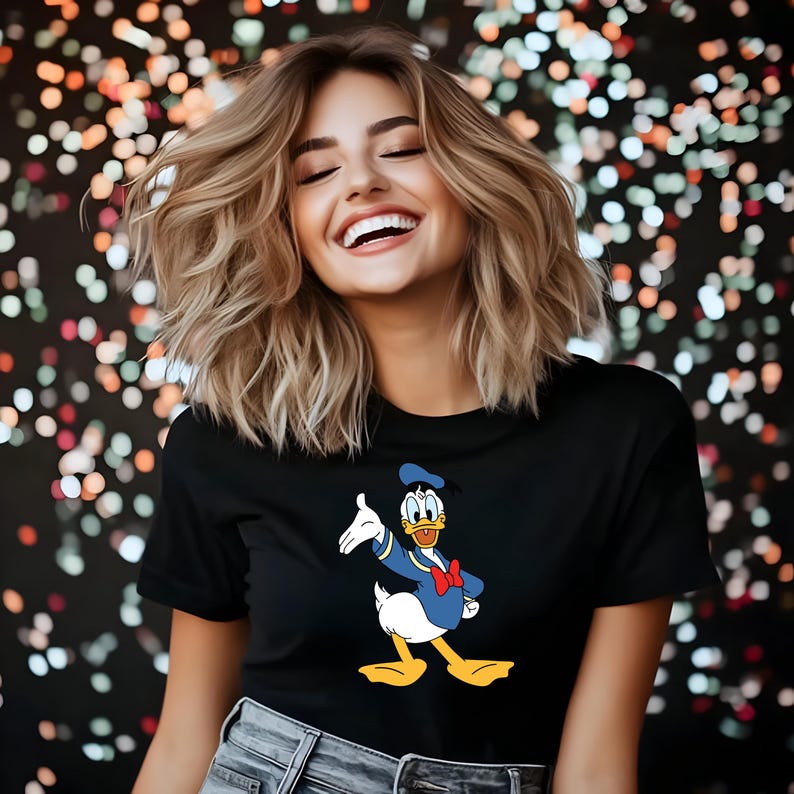 Puede incluir: Camiseta negra con una imagen de dibujos animados de Pato Donald. El personaje lleva un traje de marinero azul, una pajarita roja y un gorro de marinero. El fondo es un bokeh de luces de colores.